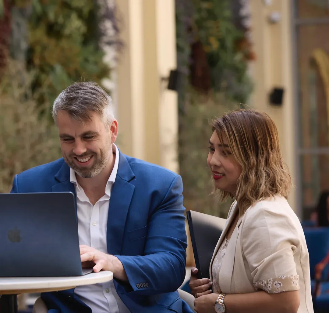 Two colleagues smiling at a laptop at an outdoor cafe table