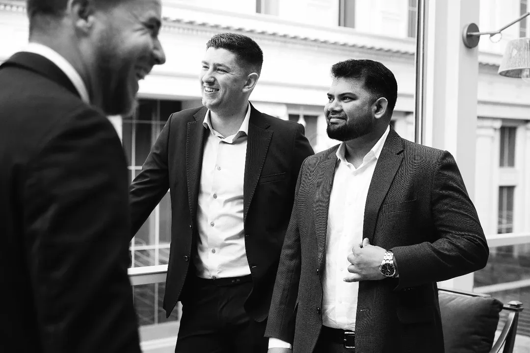 Three smiling men in suits talking beside a window, in black and white