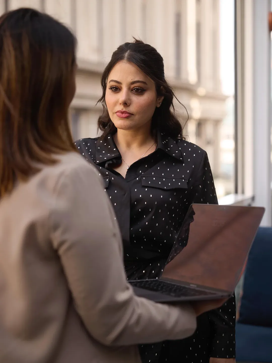 Woman in a dark spotted blouse listening to a colleague holding a laptop