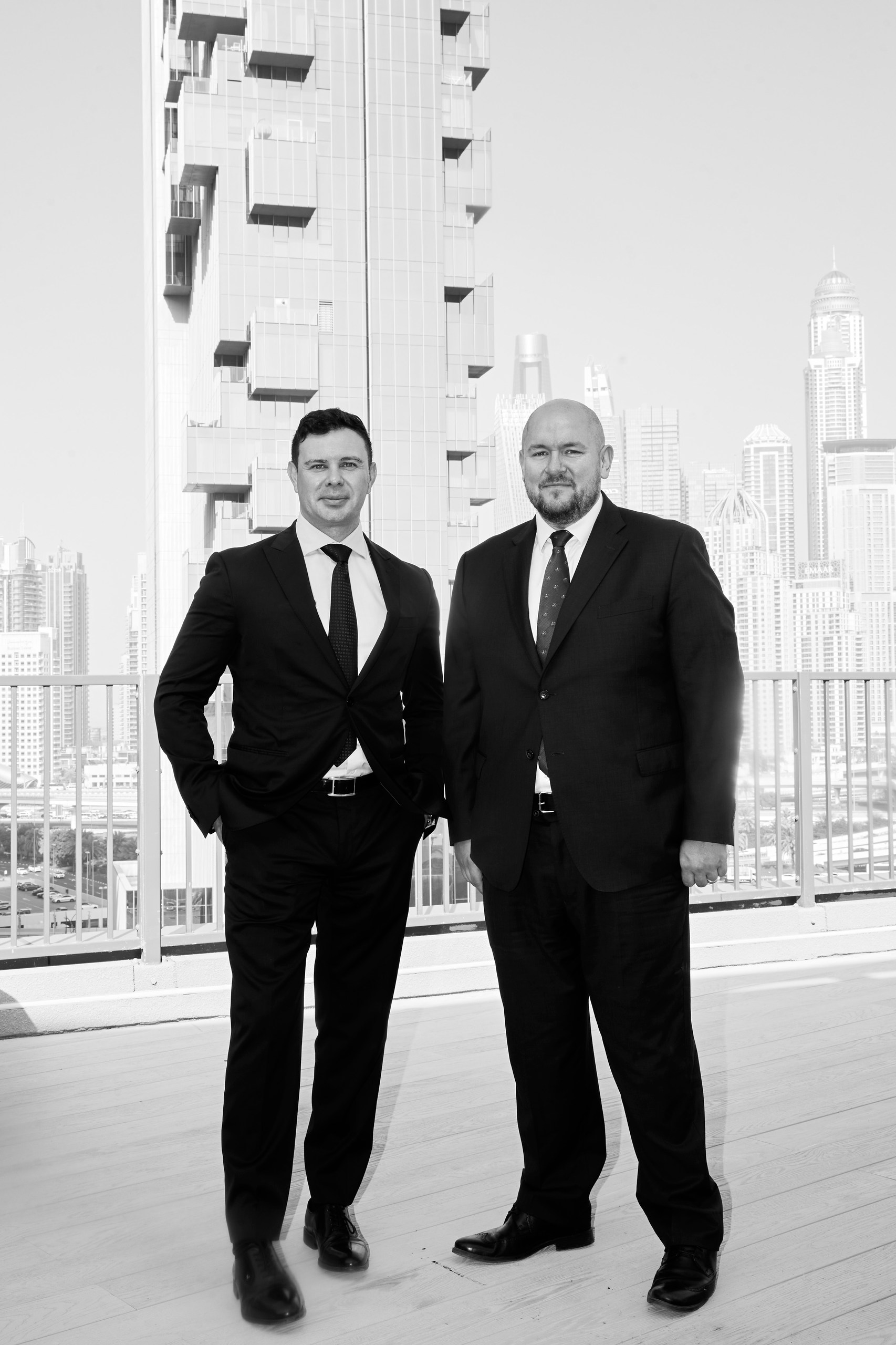 Two men in dark suits standing on a rooftop terrace with high-rise towers behind